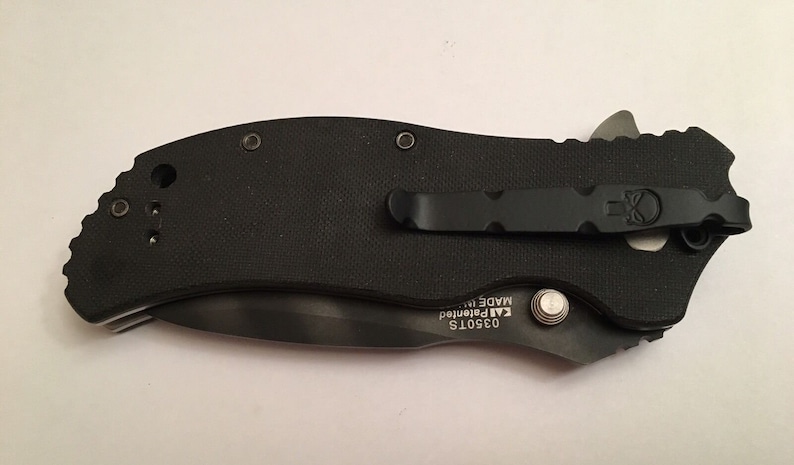 Pu&ograve; includere: Coltello pieghevole nero con un fermaglio da tasca nero e un logo a forma di teschio. Il coltello ha un'impugnatura nera e una lama argentata. La lama &egrave; parzialmente aperta e il coltello &egrave; chiuso. Il coltello ha il testo "03501S KAI Patented MADE IN" sul lato.