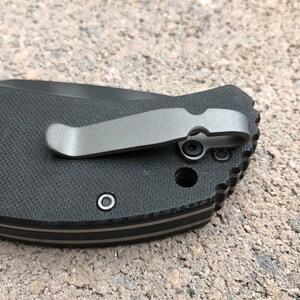 May include: Close-up of a black pocket knife with a silver pocket clip. The knife has a textured black handle and a silver pocket clip.