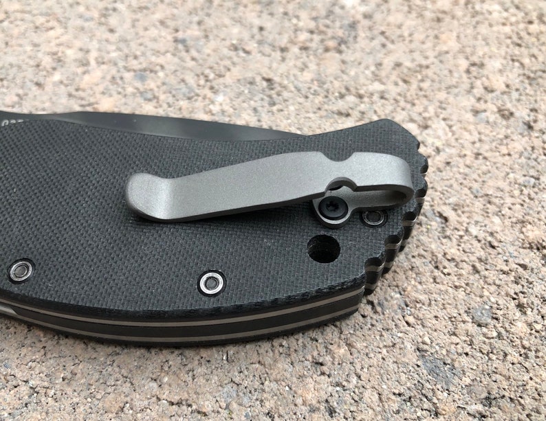May include: Close-up of a black pocket knife with a silver pocket clip. The knife has a textured black handle and a silver pocket clip.