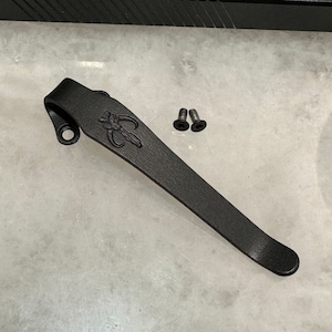 May include: Black metal pocket clip with a Mandalorian symbol engraved on it. Two small black screws are included.