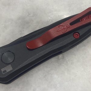 May include: A black pocket knife with a red pocket clip. The knife has a textured handle and a silver pocket clip.