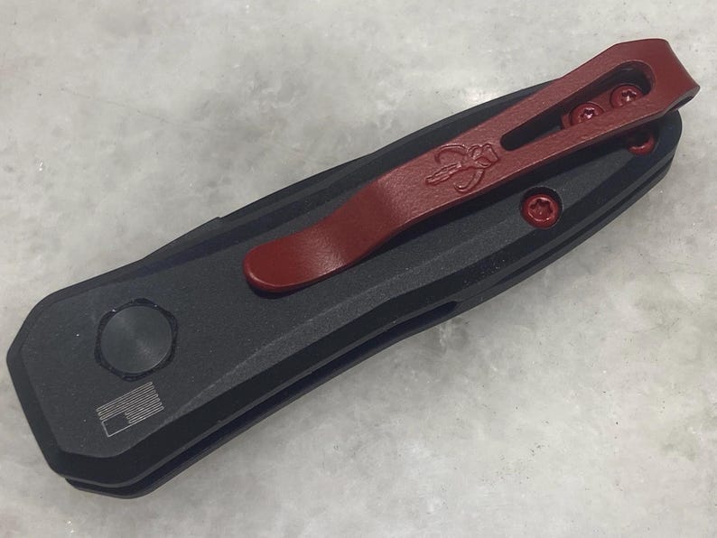 May include: A black pocket knife with a red pocket clip. The knife has a textured handle and a silver pocket clip.