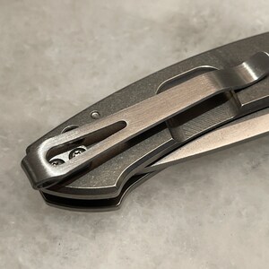 May include: A close-up of a silver pocket knife with a clip. The clip is attached to the knife with two small screws.