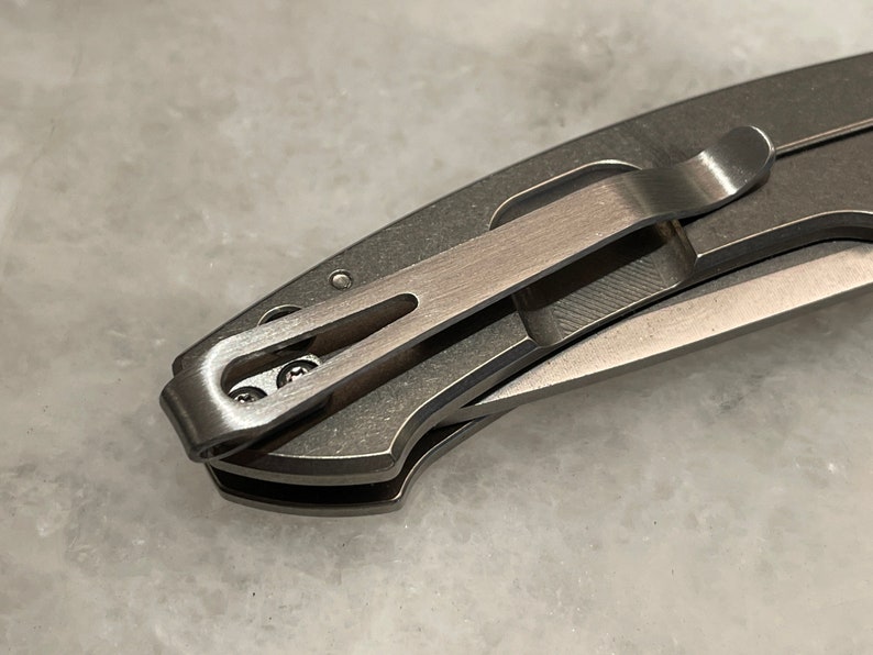 May include: A close-up of a silver pocket knife with a clip. The clip is attached to the knife with two small screws.