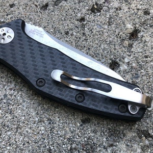 May include: A black and silver pocket knife with a carbon fiber handle and a pocket clip. The knife has a partially serrated blade and the text "KAI USA MADE IN USA" is printed on the handle.