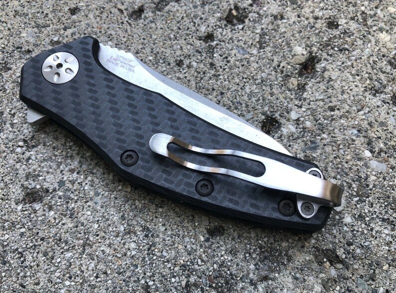 May include: A black and silver pocket knife with a carbon fiber handle and a pocket clip. The knife has a partially serrated blade and the text "KAI USA MADE IN USA" is printed on the handle.