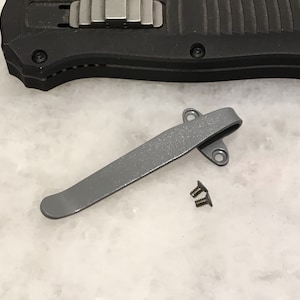 Może przedstawiać: Srebrny metalowy klips do kieszeni z dwoma małymi śrubami. Klips jest przeznaczony do mocowania do noża kieszonkowego lub innego narzędzia.