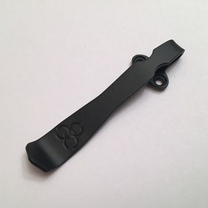 May include: Black metal pocket clip with a three-leaf clover design. The clip has two holes for attaching to a pocket or belt.