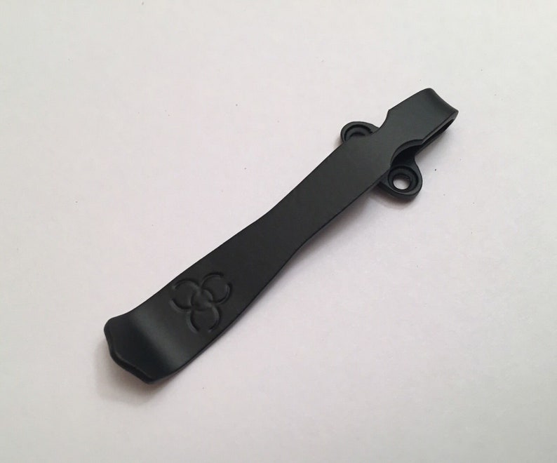May include: Black metal pocket clip with a three-leaf clover design. The clip has two holes for attaching to a pocket or belt.