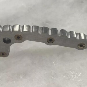 May include: A silver metal object with a textured surface and four brass-colored screws. The object has a curved shape and a series of ridges along the top.