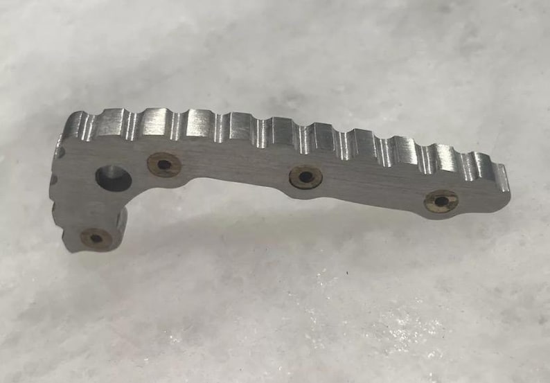 May include: A silver metal object with a textured surface and four brass-colored screws. The object has a curved shape and a series of ridges along the top.