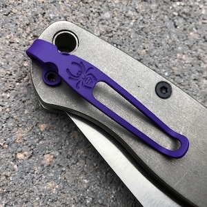May include: A purple pocket clip with a spider logo attached to a silver pocket knife.