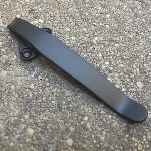 May include: A matte black metal pocket clip featuring a curved, elongated design with a wider base section that includes two circular screw holes for attachment. The surface of the clip is smooth and uniform, showing a solid dark finish throughout.