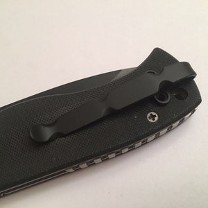 May include: A black pocket knife with a textured handle and a black pocket clip. The clip is attached to the handle with two screws.