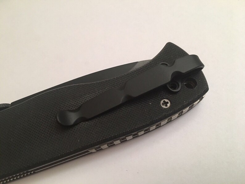 May include: A black pocket knife with a textured handle and a black pocket clip. The clip is attached to the handle with two screws.