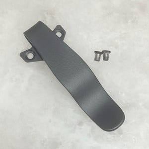 May include: A black pocket clip with two silver screws. The clip is made of metal and has a curved design. It features two holes for mounting and a textured surface.