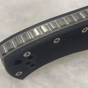 May include: Close-up of a black pocket knife with a textured handle and a silver blade. The handle has a textured pattern and four screws.