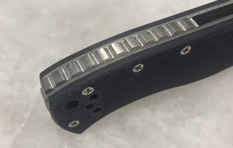 May include: Close-up of a black pocket knife with a textured handle and a silver blade. The handle has a textured pattern and four screws.