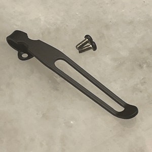 Op de afbeelding: Zwarte metalen zakclip met twee schroeven. De clip heeft een afgerond uiteinde en een klein gaatje in de buurt van de bovenkant.