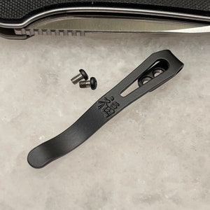 Könnte beinhalten: Schwarzer Metall-Taschenclip mit einem gravierten japanischen Schriftzeichen. Zwei kleine schwarze Schrauben sind im Lieferumfang enthalten.