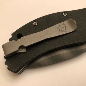 Puede incluir: Un cuchillo de bolsillo negro con un clip de bolsillo plateado. El clip tiene un peque&ntilde;o logotipo. El cuchillo tiene el texto "03501S KAI Patented MADE IN JAPAN" en el lateral.
