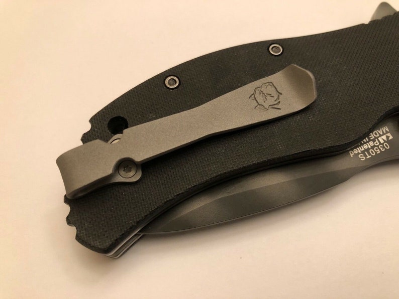 Puede incluir: Un cuchillo de bolsillo negro con un clip de bolsillo plateado. El clip tiene un peque&ntilde;o logotipo. El cuchillo tiene el texto "03501S KAI Patented MADE IN JAPAN" en el lateral.