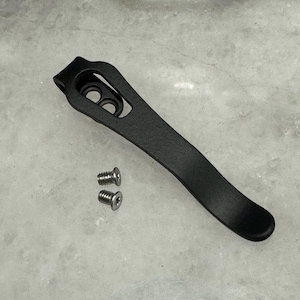 May include: Black metal pocket clip with two screws. The clip has a curved shape and a hole in the top.