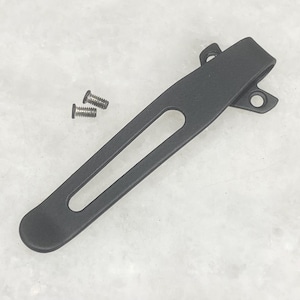 May include: A black pocket clip with a long, slender design and a curved end. Two small silver screws are included. The clip has a rectangular cutout and two holes for attachment.