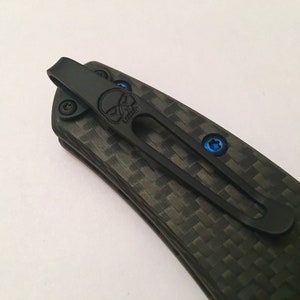 Puede incluir: Cuchillo de bolsillo negro con mango de fibra de carbono y un clip en forma de calavera. El clip est&aacute; sujeto con tornillos azules.