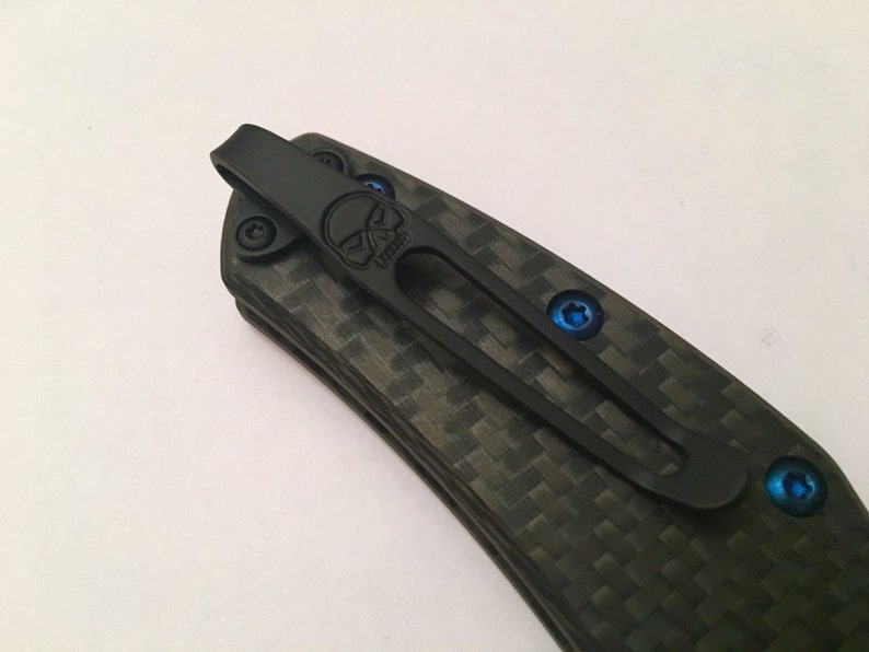 Puede incluir: Cuchillo de bolsillo negro con mango de fibra de carbono y un clip en forma de calavera. El clip est&aacute; sujeto con tornillos azules.