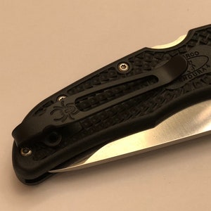 May include: Black pocket knife with a black clip and a textured black handle. The blade is partially visible and has a silver finish. The handle has the text "ERGO" and "ENDURA4" on it.