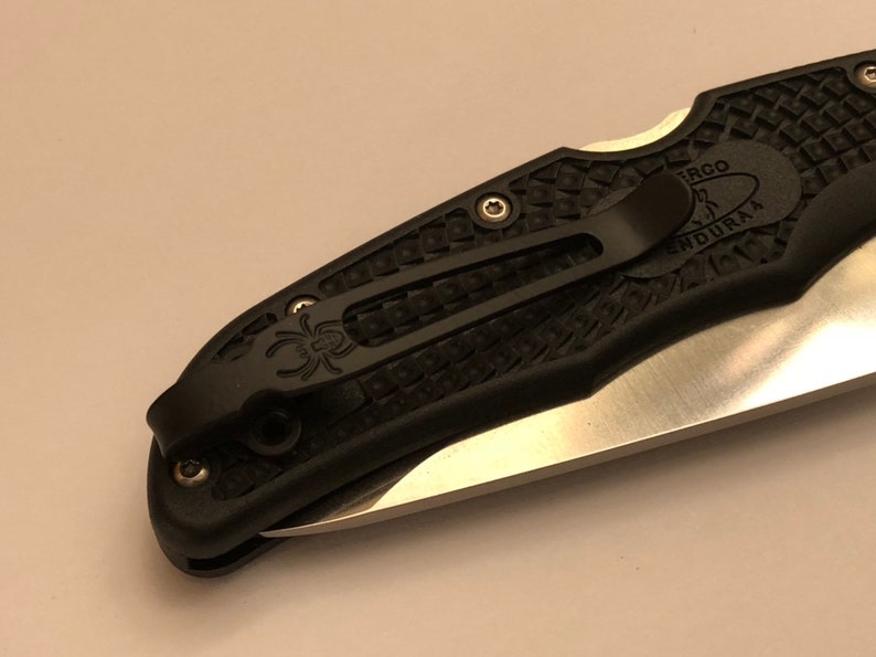 May include: Black pocket knife with a black clip and a textured black handle. The blade is partially visible and has a silver finish. The handle has the text "ERGO" and "ENDURA4" on it.