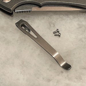 May include: A silver pocket knife clip with two screws. The clip is curved and has a hole in the center.