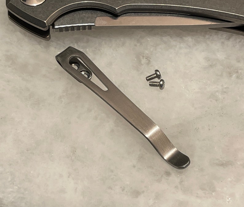 May include: A silver pocket knife clip with two screws. The clip is curved and has a hole in the center.