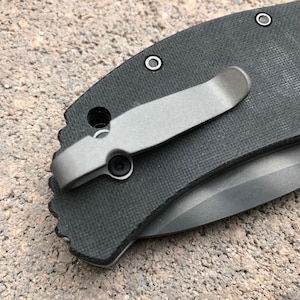 May include: A close-up of a black pocket knife with a silver clip. The knife has a textured handle and a silver blade.