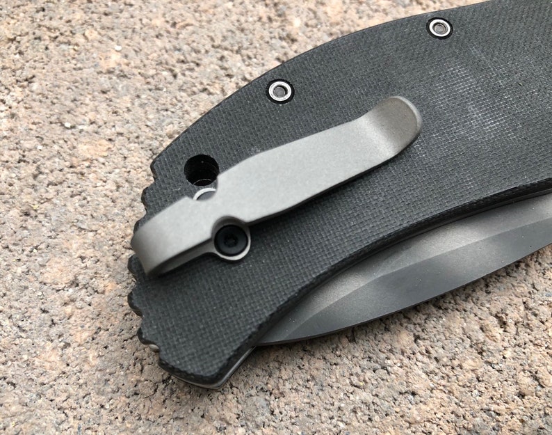May include: A close-up of a black pocket knife with a silver clip. The knife has a textured handle and a silver blade.