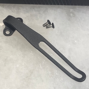 May include: Black metal pocket clip with two screws. The clip has a smooth, matte finish and a rounded, open loop at the end.