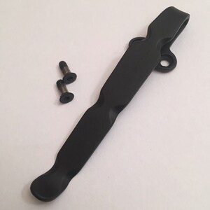 May include: Black metal pocket clip with two small screws. The clip has a curved design and a hole at the top.