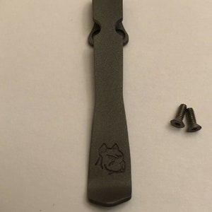 Puede incluir: Un clip de bolsillo de metal gris oscuro con un dise&ntilde;o de cabeza de perro grabado en la parte delantera. Se incluyen dos peque&ntilde;os tornillos plateados.