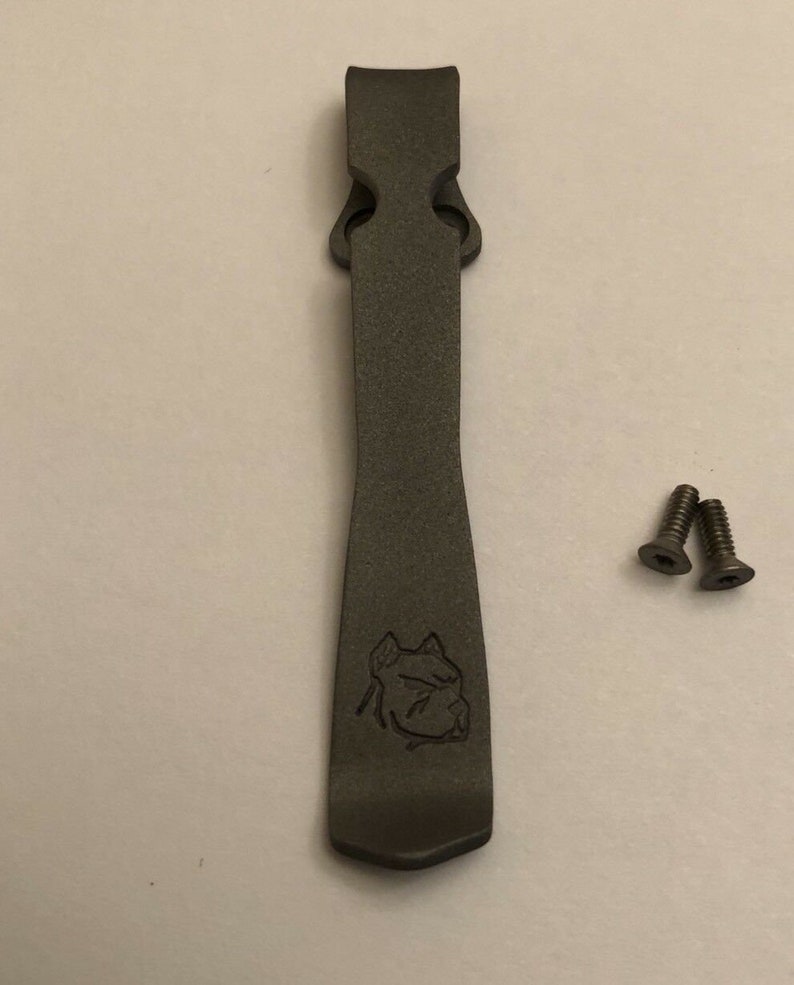 Puede incluir: Un clip de bolsillo de metal gris oscuro con un dise&ntilde;o de cabeza de perro grabado en la parte delantera. Se incluyen dos peque&ntilde;os tornillos plateados.