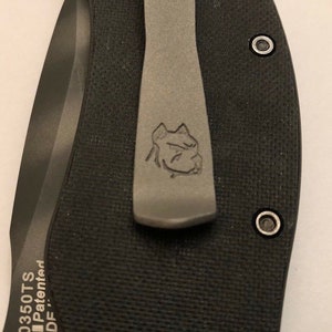 Puede incluir: Primer plano de una navaja de bolsillo negra con un clip de bolsillo plateado. El clip de bolsillo tiene un logotipo de cabeza de perro grabado. La navaja tiene el texto "0350TS AIP Patented ADF" grabado en el mango.