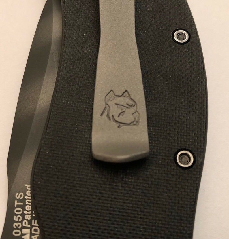 Puede incluir: Primer plano de una navaja de bolsillo negra con un clip de bolsillo plateado. El clip de bolsillo tiene un logotipo de cabeza de perro grabado. La navaja tiene el texto "0350TS AIP Patented ADF" grabado en el mango.