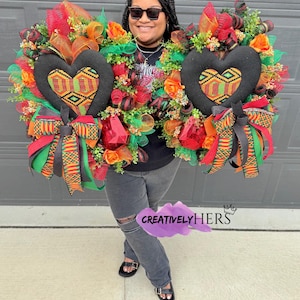 May include: Two heart-shaped wreaths with black fabric centers featuring a colorful geometric pattern. The wreaths are adorned with red, orange, and green flowers, ribbons, and mesh. The text "CREATIVELYHERS" is visible.