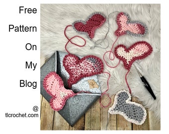 May include: Five crocheted hearts in various shades of pink, red, and gray, with a crochet hook and yarn on a fuzzy white surface. The text "Free Pattern On My Blog @ tlcrochet.com" is visible in the image.