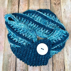 May include: A teal blue crocheted infinity scarf with a white button. The scarf is made with a textured stitch and has a gradient of blue colors.