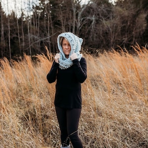 May include: A woman wearing a gray hooded cowl with black bobbles. She is standing in a field of tall, dry grass.