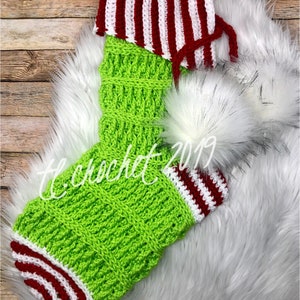 Puede incluir: Un calcetín de Navidad de crochet a rayas verdes y blancas con un puño a rayas rojas y blancas. El calcetín está decorado con un pompón de piel sintética blanca.