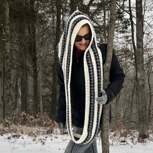 May include: A black and white striped hooded scarf with a crocheted design. The scarf is being worn by a person in a snowy woodland setting.