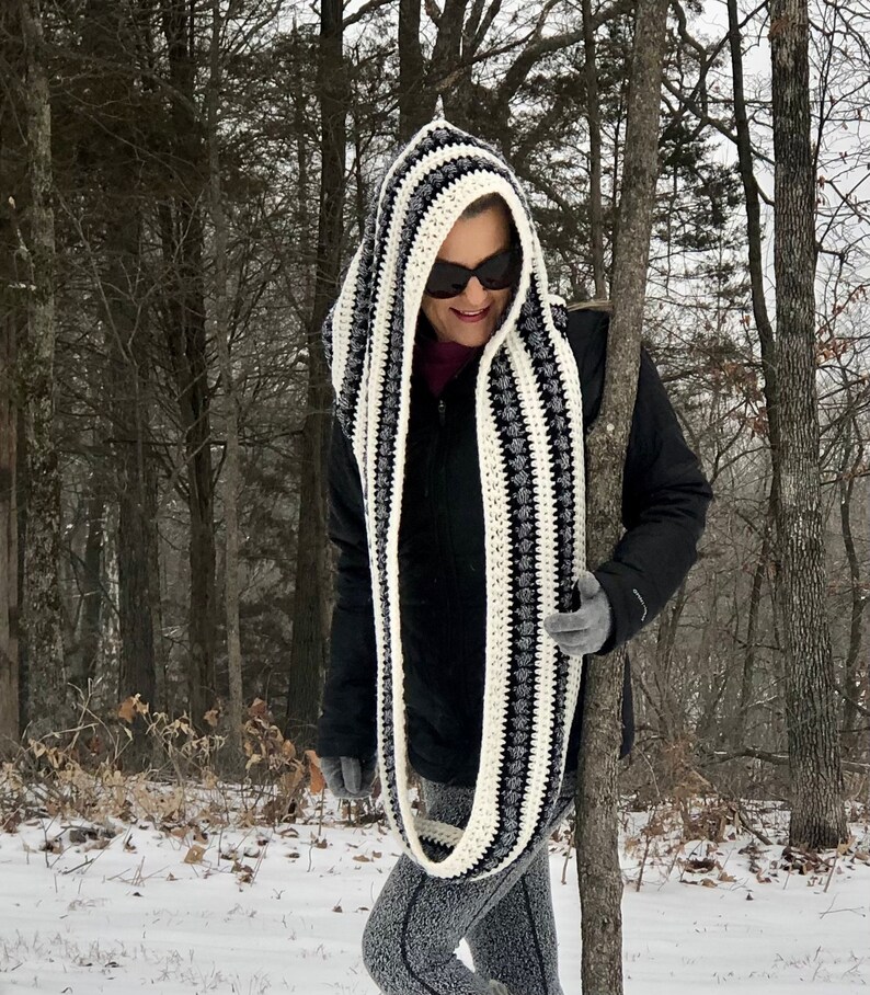 May include: A black and white striped hooded scarf with a crocheted design. The scarf is being worn by a person in a snowy woodland setting.