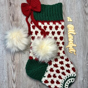 May include: A green, red, and white crocheted Christmas stocking with a red bow and two white fur pom-poms. The stocking has a heart pattern and is decorated with a wooden sign that says "Calicooft".
